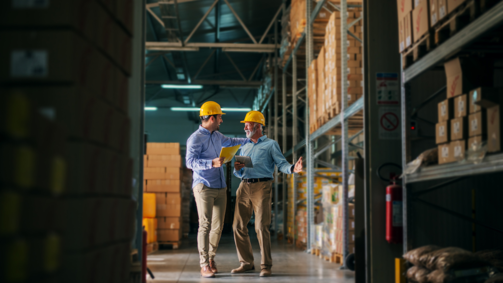 Two men in a warehouse wearing hard hats have a conversation while holding a clipboard